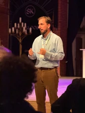 A man in a light blue shirt gestures while speaking to an audience in a dimly lit venue with a stage and candelabra in the background.