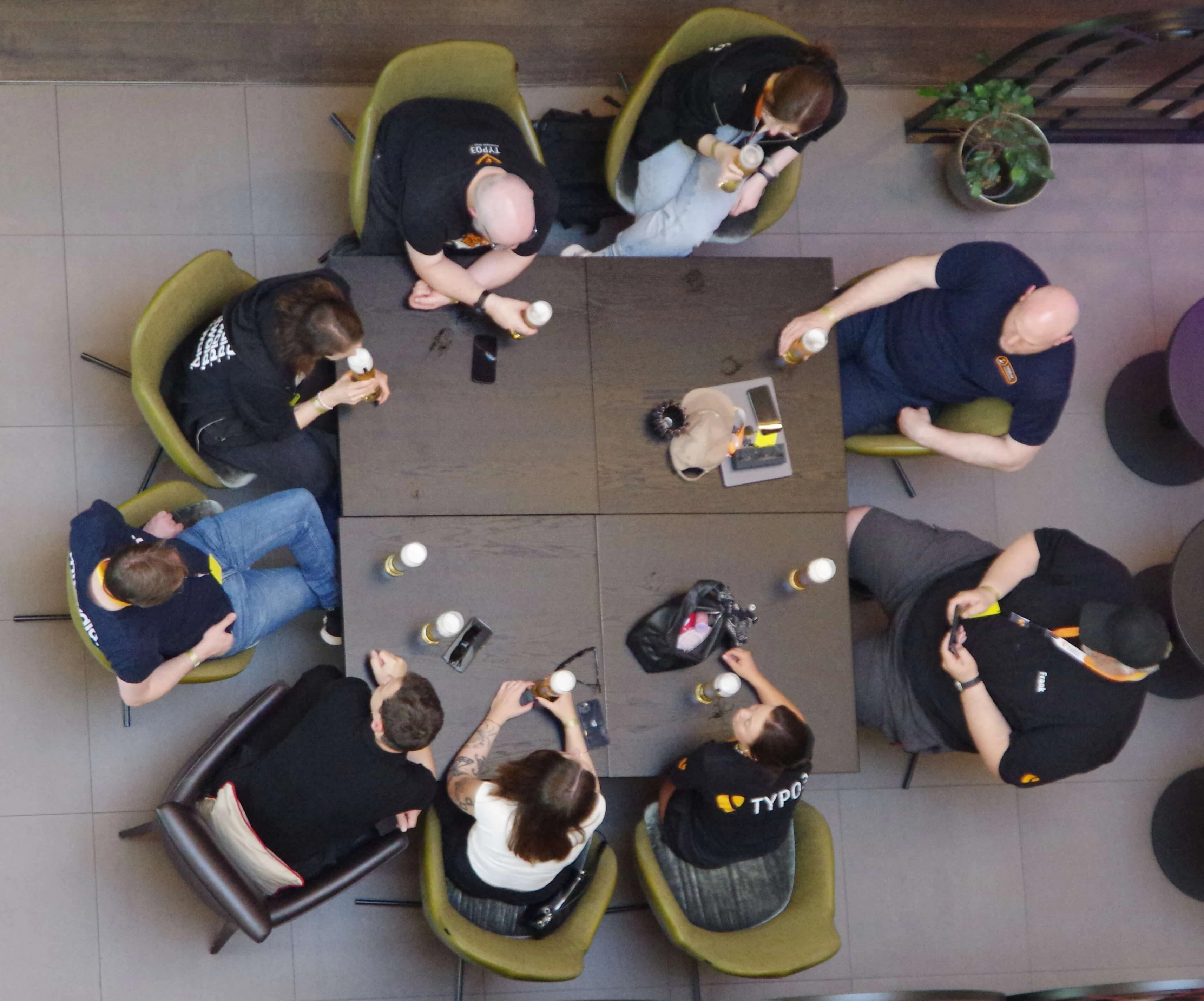 A group of eight people seated around a square table, engaged in conversation, with drinks and snacks on the table.