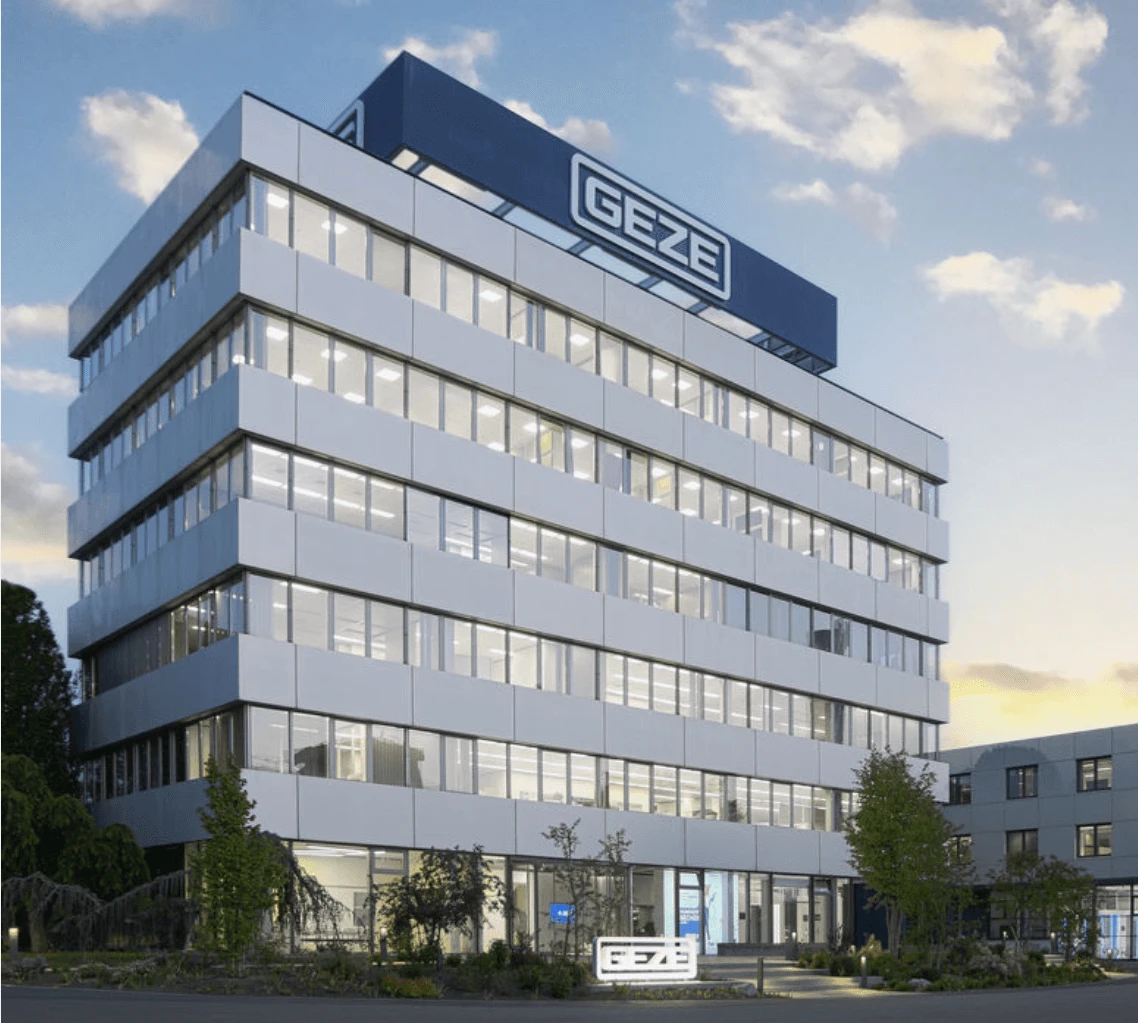 Modern office building with the GEZE logo prominently displayed on top, surrounded by greenery and a secondary structure nearby.