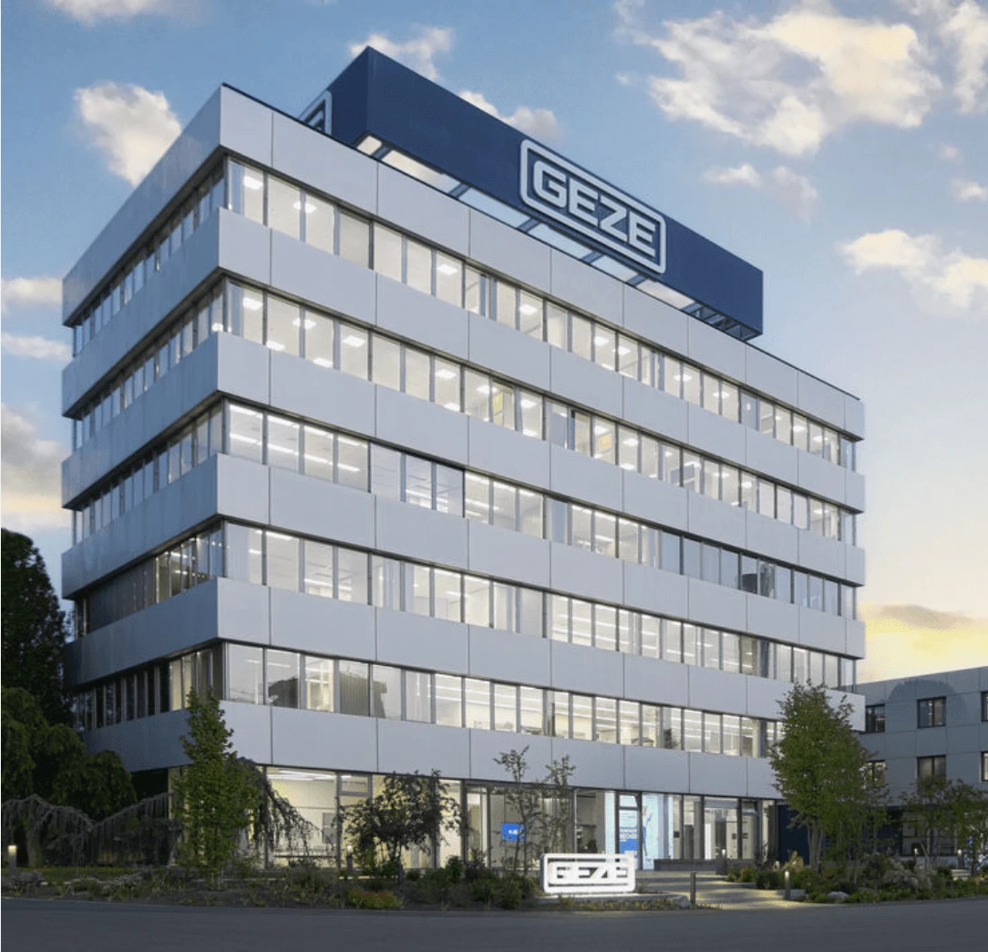 Modernes Bürogebäude mit dem deutlich sichtbaren GEZE-Logo auf dem Dach, umgeben von Grünflächen und einem angrenzenden Nebengebäude.