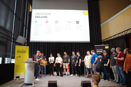 A group of people stands on stage during an event, with a presentation screen displaying "ORGATEAM DELUXE" and sponsor logos in the background.
