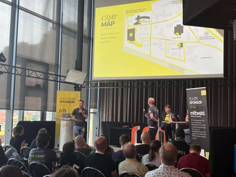 Zwei Referenten präsentieren auf einer Konferenz, mit einer großen Karte, die hinter ihnen angezeigt wird. Die Teilnehmer sitzen im Vordergrund und sind in die Sitzung vertieft.