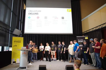 Eine Gruppe von Menschen steht während einer Veranstaltung auf der Bühne, im Hintergrund ist ein Präsentationsbildschirm mit der Aufschrift "ORGATEAM DELUXE" und den Logos der Sponsoren zu sehen.