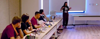 A speaker presents to an audience seated at a long table, with various drinks and notebooks in front of them.
