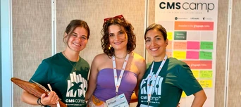 Three women pose together, wearing CMS camp t-shirts, with a colorful bulletin board in the background displaying various notes and information.