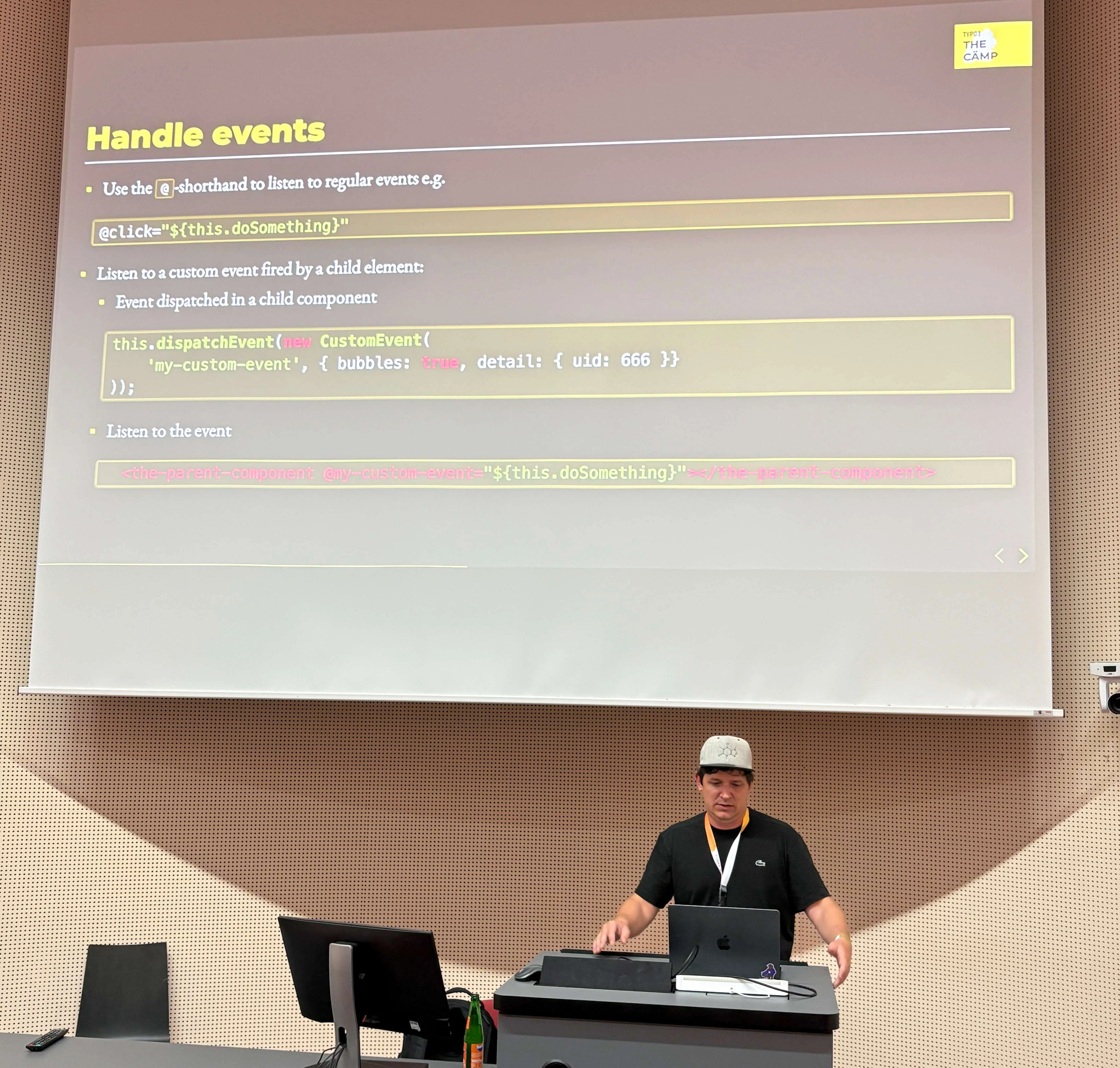 A speaker presents on handling events in programming, with code examples displayed on a large screen behind him.