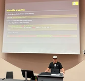 Ein Redner präsentiert, wie man Ereignisse in der Programmierung behandelt, während Codebeispiele auf einer großen Leinwand hinter ihm angezeigt werden.