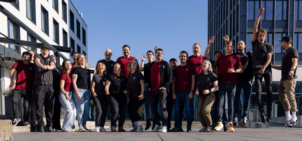 A diverse group of people stands together outdoors, wearing black and red shirts, with some posing energetically in front of modern buildings.