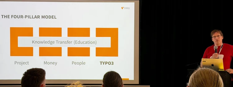 Ein Präsentator steht vor einer Folie, die "Das Vier-Säulen-Modell" zeigt und hebt die Bereiche Projekt, Geld, Menschen und Wissensaustausch (Bildung) für TYPO3 hervor.