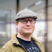 Man wearing a gray cap and glasses, dressed in a black shirt and green jacket, standing in a well-lit indoor environment.