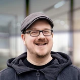 Man with glasses and a flat cap smiles, wearing a dark hoodie, against a blurred indoor background.