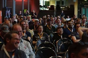 Ein diverses Publikum von Menschen, die in Reihen sitzen, lächeln und lachen während einer Präsentation oder Veranstaltung, wobei einige Lanyards tragen.