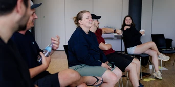 A group of five people seated in a casual setting, engaged in conversation, with some holding drinks and smiling.