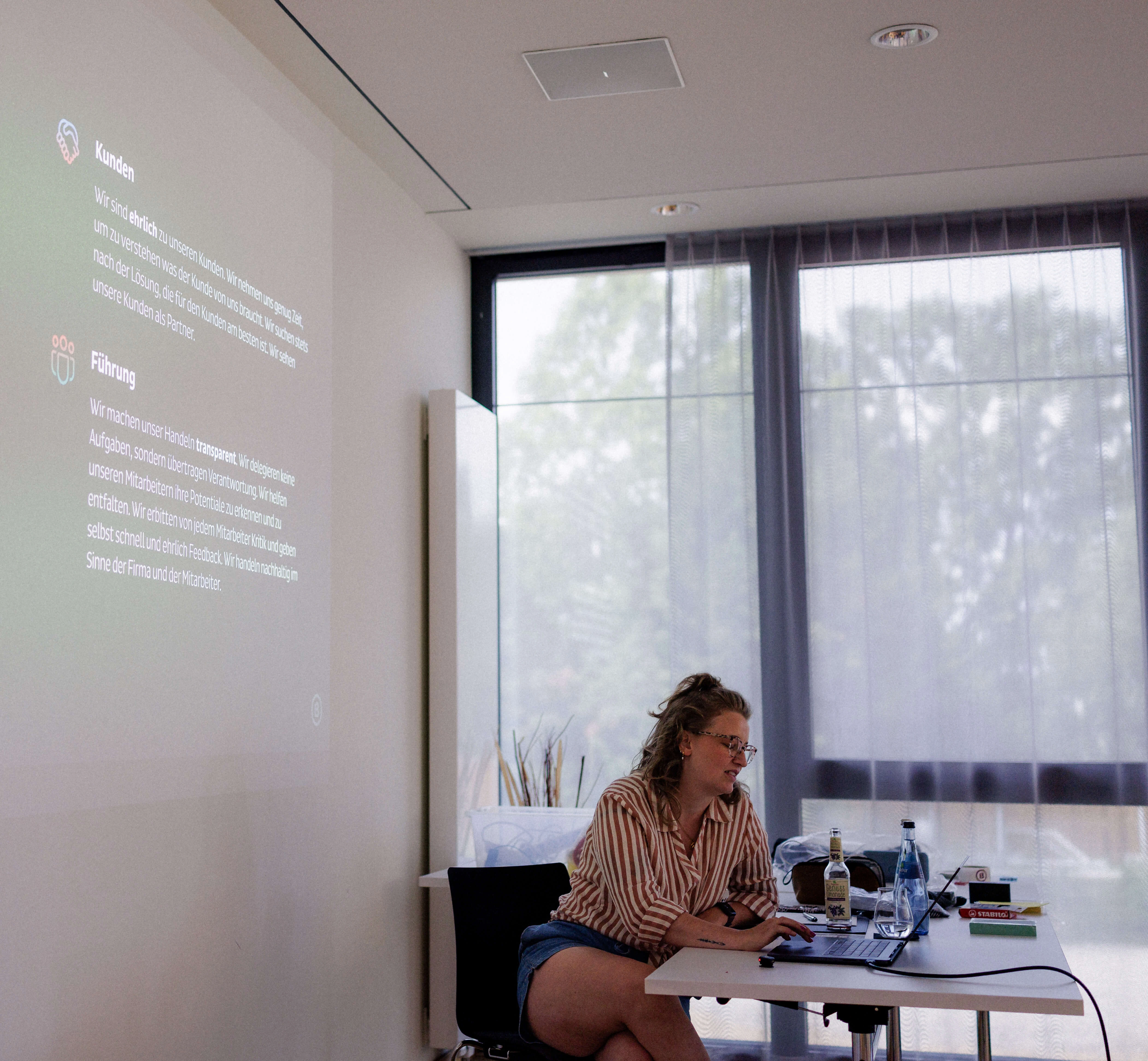 A woman with curly hair sits at a table with a laptop, engaged in a presentation. A projector displays text on the wall beside her.