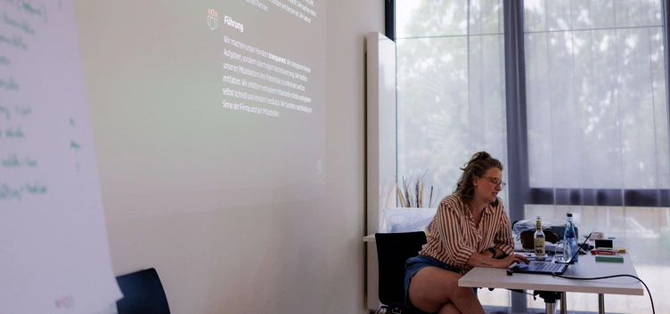 A woman with curly hair sits at a table with a laptop, engaged in a presentation. A projector displays text on the wall beside her.