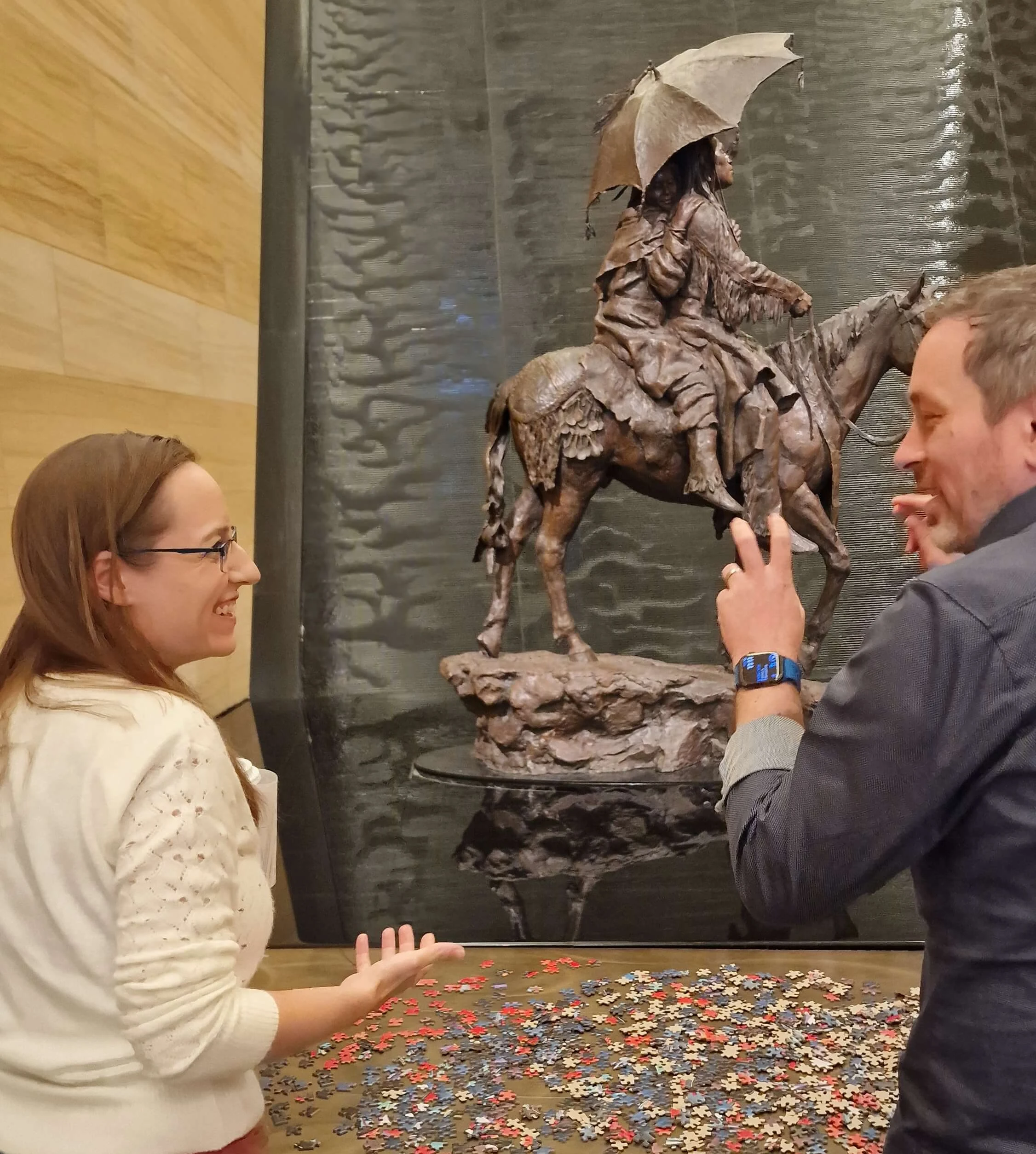 Two people engage in conversation near a large puzzle spread out on a table, with a statue of a mounted figure holding an umbrella in the background.