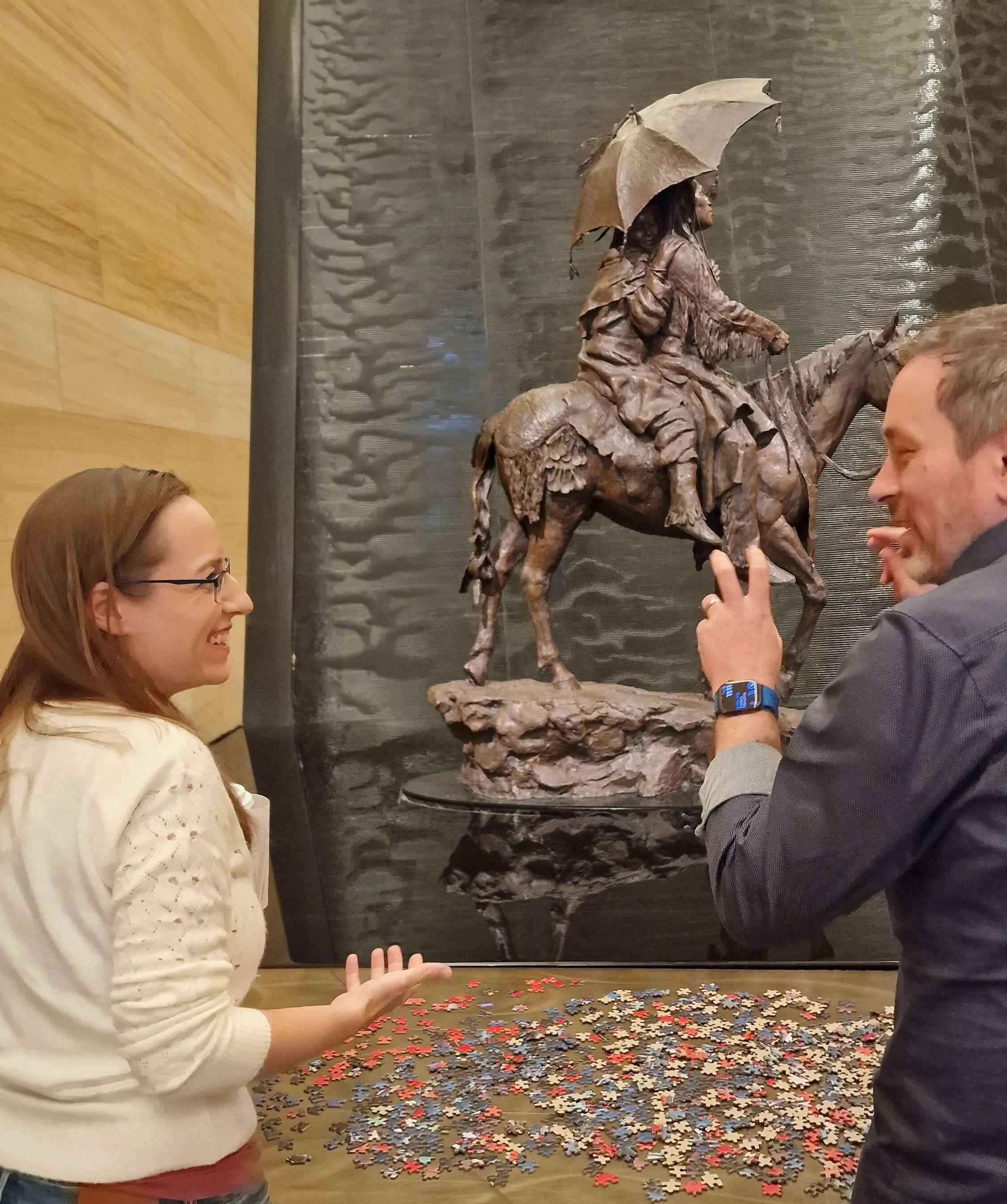 Zwei Personen führen ein Gespräch in der Nähe eines großen Puzzles, das auf einem Tisch ausgebreitet ist, während im Hintergrund eine Statue einer berittenen Figur mit einem Regenschirm steht.