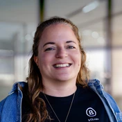Smiling woman with long hair wearing a black shirt and denim jacket, standing in a well-lit indoor space.