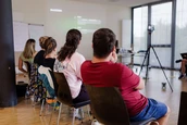 Eine Gruppe von Menschen sitzt in Stühlen und blickt auf eine Projektionsleinwand, die Text anzeigt, während eine Kamera in der Nähe aufgestellt ist.