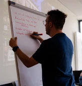 A person writes on a flip chart during a meeting, while another individual works on a laptop in the background.