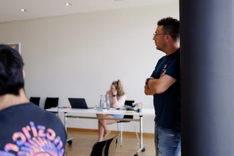 A man stands with arms crossed, observing a woman seated at a table with laptops and water bottles in a modern classroom setting.