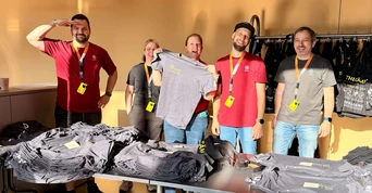Gruppe von fünf Personen, die hinter einem Tisch mit gestapelten grauen T-Shirts stehen, lächeln und Namensschilder tragen, in einer gut beleuchteten Innenumgebung.