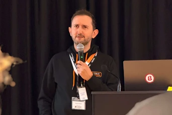 A speaker stands at a podium, holding a microphone, while presenting to an audience. A laptop is visible on the podium.