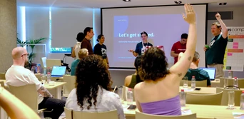 A group of people in a conference room, with one person raising their hand while others engage in discussion. A presentation screen displays "Let's get started.