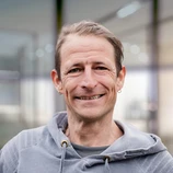 Mann mit schulterlangen Haaren, der einen grauen Hoodie trägt und in einem Innenraum mit unscharfen Hintergrundelementen lächelt.