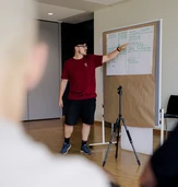 A man in a red shirt presents to an audience, pointing at a large paper on an easel with notes and diagrams.