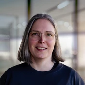 Woman with shoulder-length gray hair and glasses, wearing a black shirt, smiling in a modern indoor setting.