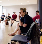 A woman sits on a chair, smiling and engaged, while a group of people are seated in a circle in a well-lit room.