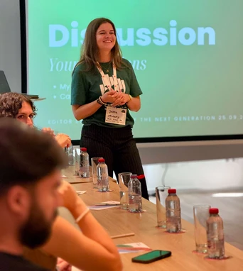 A woman in a green shirt stands at the front of a conference room, leading a discussion while attendees listen at a table.