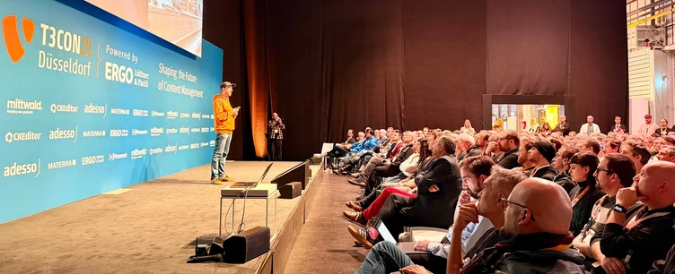 Ein Redner in einem orangefarbenen Hemd präsentiert auf der Bühne einer Konferenz, während das Publikum aufmerksam vor einem markenbezogenen Hintergrund zuhört.