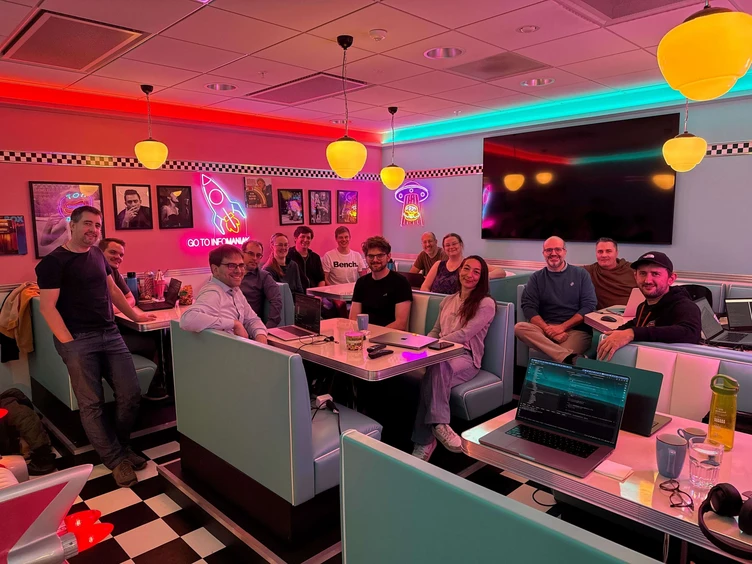 Gruppe von Menschen, die in einem Retro-Diner sitzen und sich unterhalten, umgeben von bunten Neonlichtern und einem großen Bildschirm im Hintergrund.