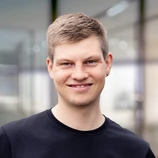 Young man with short light hair and earrings, smiling while wearing a black shirt, in a modern office environment.