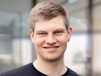 Junger Mann mit kurzen, hellen Haaren und Ohrringen, der in einem modernen Büroumfeld lächelt und ein schwarzes Hemd trägt.