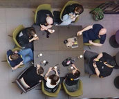 Eine Gruppe von acht Personen sitzt um einen quadratischen Tisch, führt ein Gespräch, während Getränke und Snacks auf dem Tisch stehen.