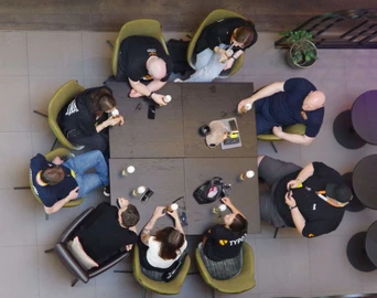 A group of eight people seated around a square table, engaged in conversation, with drinks and snacks on the table.