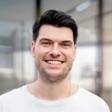 Young man with short dark hair and a slight beard, smiling while wearing a white shirt, in a modern indoor setting.