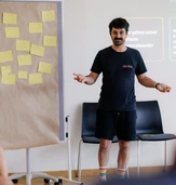 A man in a black t-shirt and shorts gestures while speaking in front of a board covered with yellow sticky notes during a workshop.