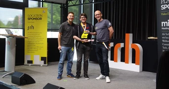 Drei Personen stehen auf der Bühne, eine hält einen Pokal, im Hintergrund ist ein Banner zu sehen, das den Standort des Sponsors der Veranstaltung anzeigt.