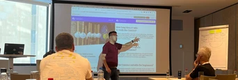 A presenter stands in front of a screen displaying a website, while an audience of seated participants listens attentively in a conference room.