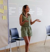 A woman in a green romper gestures while speaking in front of a presentation screen with text in German about customer relations and responsibilities.