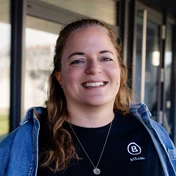 Lächelnde Frau mit langen Haaren, die ein schwarzes T-Shirt und eine Jeansjacke trägt, steht vor einem neutralen Hintergrund.