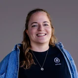 Lächelnde Frau mit langen Haaren, die ein schwarzes T-Shirt und eine Jeansjacke trägt, steht vor einem neutralen Hintergrund.