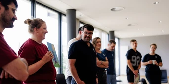 A group of people stands in a bright room, engaged in conversation, with some holding papers and others listening attentively.