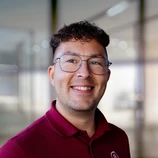 Smiling man with curly hair and glasses, wearing a red polo shirt, stands in a well-lit indoor setting.