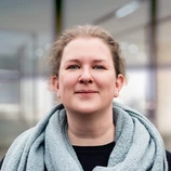 Woman with light brown hair wearing a gray scarf and black top, standing in a modern indoor setting.