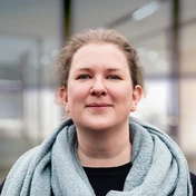 Woman with light brown hair wearing a gray scarf and black top, standing in a modern indoor setting.